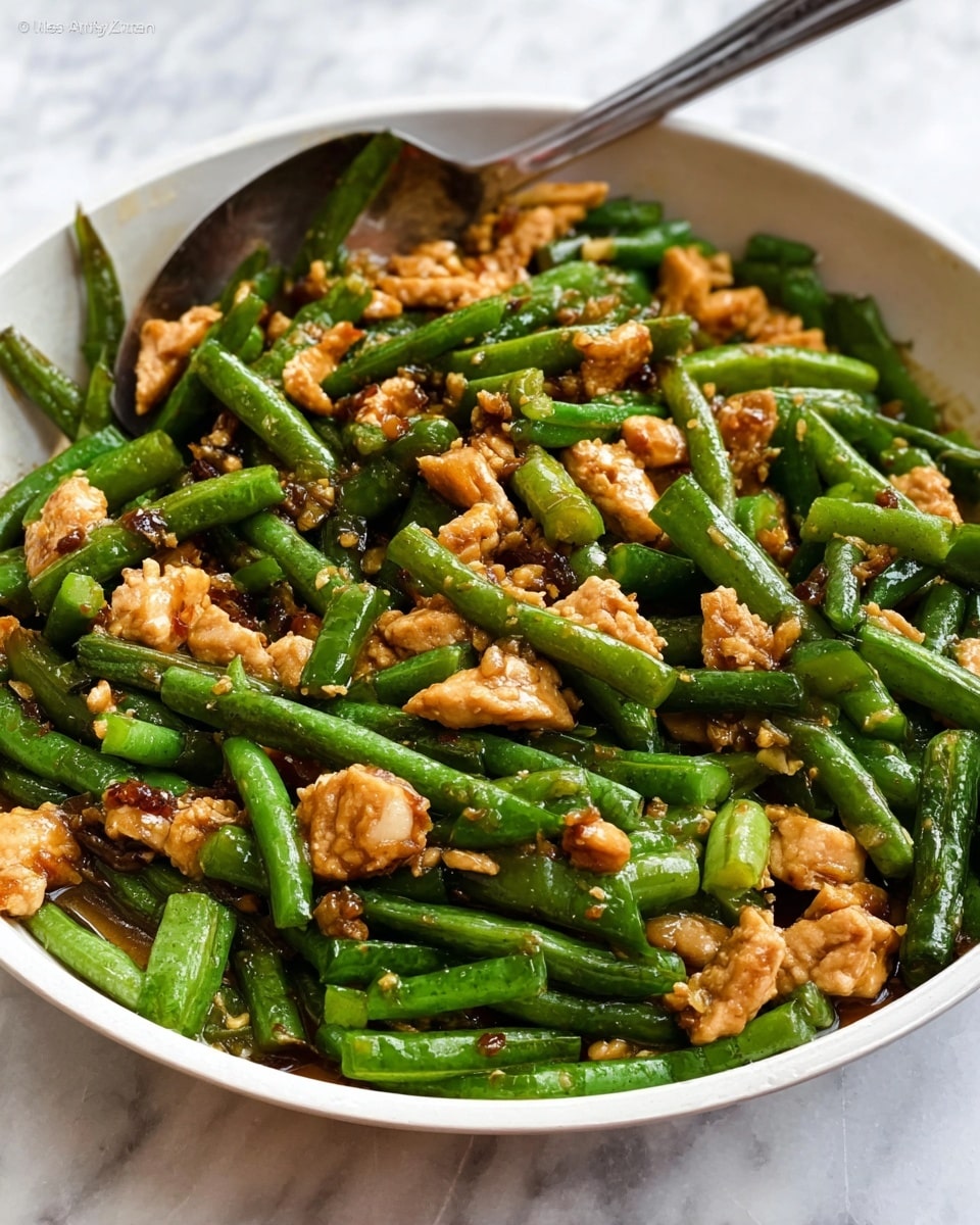 The image shows a close-up of a dish in a simple white bowl. The bowl is full of chopped green beans and pieces of light brown cooked tempeh mixed together. The green beans are cut into short segments and have a fresh green color, while the tempeh pieces have a slightly crispy texture with a golden brown color. The food is glistening slightly, indicating it is cooked in oil or sauce. The bowl sits on a white marbled surface, and there is a silver serving spoon partially inside the dish, ready to serve. photo taken with an iphone --ar 4:5 --v 7