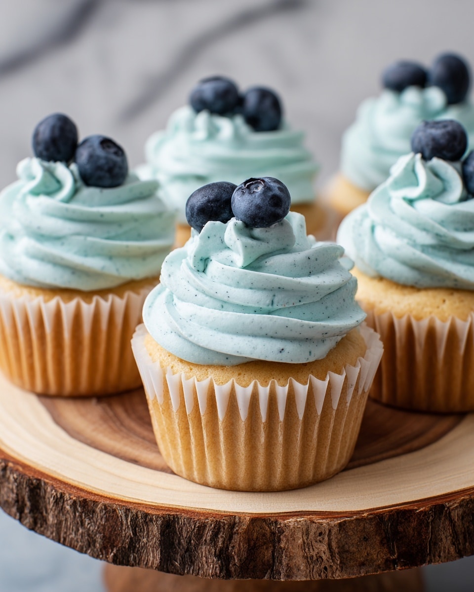 A close-up of five cupcakes arranged on a round wooden slab, each cupcake has a light golden brown base with white paper liners, topped with a thick swirl of light blue frosting that has a smooth and creamy texture with tiny dark specks. On top of each frosting swirl, there are two fresh blueberries placed neatly. The background features a white marbled texture that adds a soft and clean look. photo taken with an iphone --ar 4:5 --v 7
