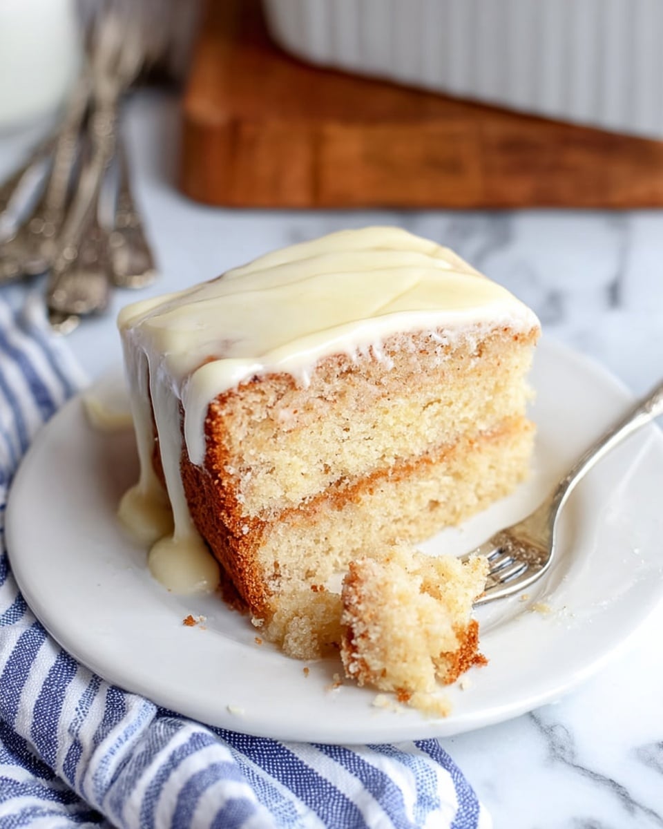 A white plate holds a thick square slice of cake with three visible layers: the base is light brown with a soft, dense texture, the middle is pale yellow and moist, and the top is coated with a creamy, white icing that drips slightly down the sides. A small piece of the cake has been cut and is on a silver fork resting on the plate, showing the light spongy inside. The plate sits on a white marbled surface next to a folded blue and white striped cloth, and the background has silver forks and a white baking dish. Photo taken with an iphone --ar 4:5 --v 7