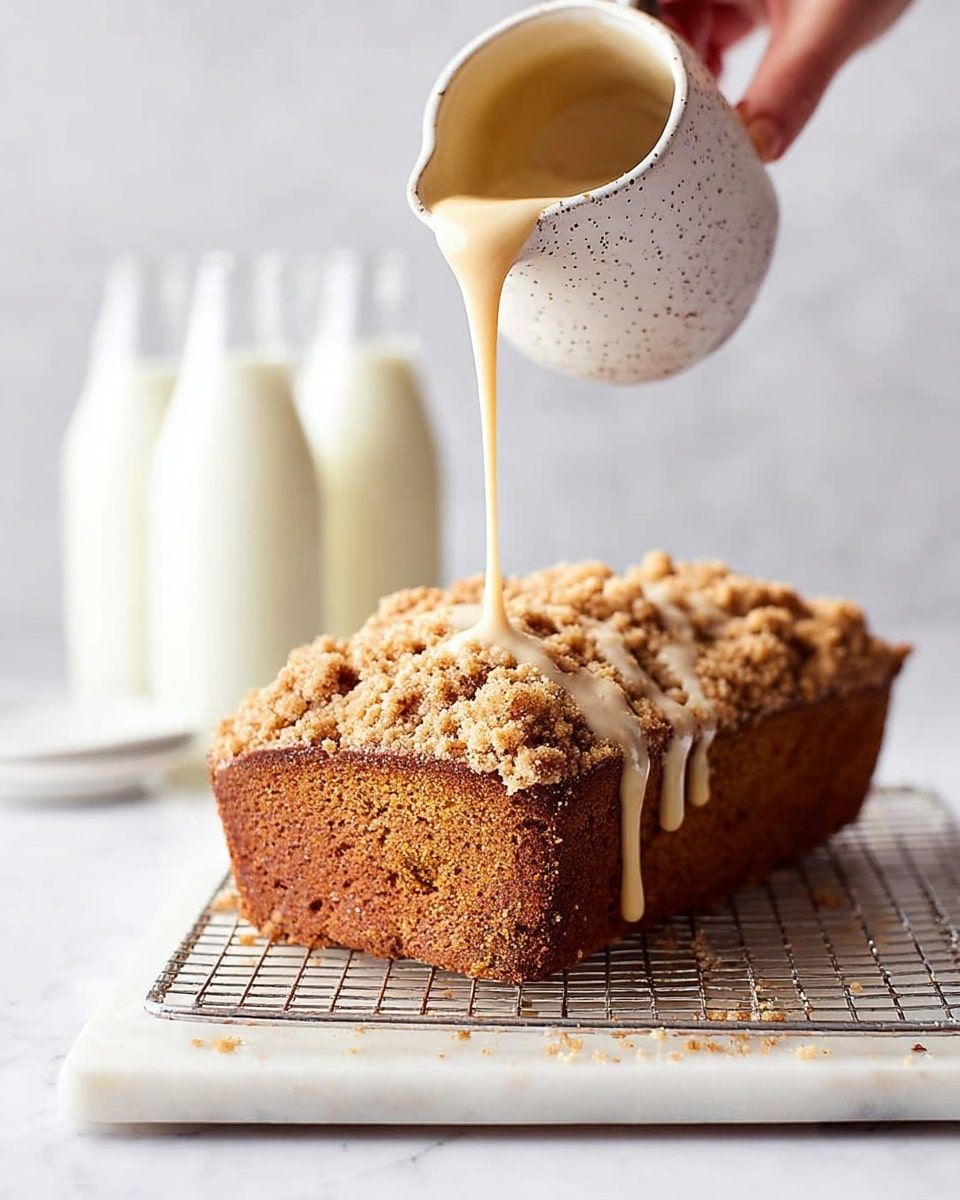 A golden brown loaf cake with a crumbly, coarse streusel topping sits on a metal cooling rack over a white marble slab, the cake's sides showing a moist texture with a darker brown crust at the base. A woman's hand pours a smooth, creamy beige glaze from a small white speckled ceramic pitcher, the glaze slowly flowing down the crumbs and sides of the cake. In the background, there are two clear bottles filled with a white liquid blurred softly against a white marbled surface. photo taken with an iphone --ar 4:5 --v 7