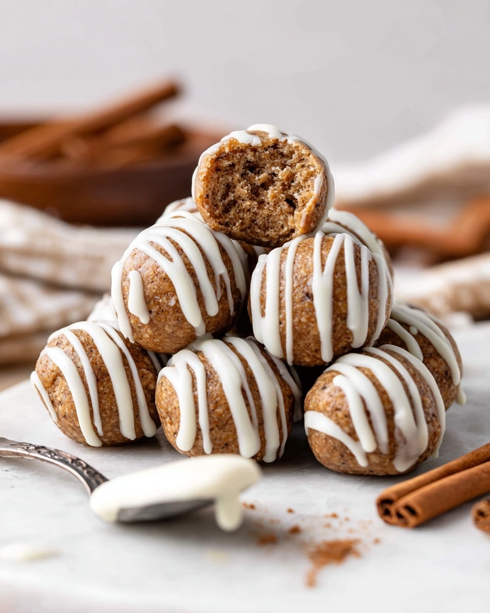 A pile of small, round brown cake balls with a soft, slightly crumbly texture is shown stacked on a white marbled surface; they are decorated with thin, white icing stripes drizzled evenly over their tops and sides, with one cake ball on top with a bite taken out, revealing a dense, moist inside with visible bits of oats or nuts; in front on the left, there is a silver spoon holding some white icing, and in the blurred background, cinnamon sticks lay scattered on the same white marbled surface. photo taken with an iphone --ar 4:5 --v 7