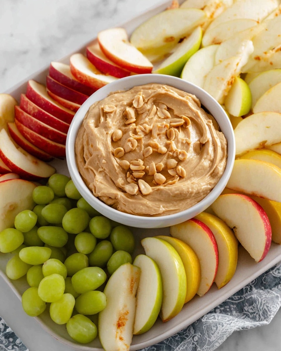 A white bowl filled with creamy peanut butter dip topped with small peanut pieces sits at the center of a white platter. Surrounding the bowl are neatly arranged apple slices in three types: red, green, and yellow, placed in groups around the bowl to create a colorful contrast. The apple slices have a smooth, shiny texture, and the peanut butter dip has a slightly swirled, thick texture with crunchy bits on top. The background is a white marbled texture with part of a patterned cloth visible on the side. photo taken with an iphone --ar 4:5 --v 7