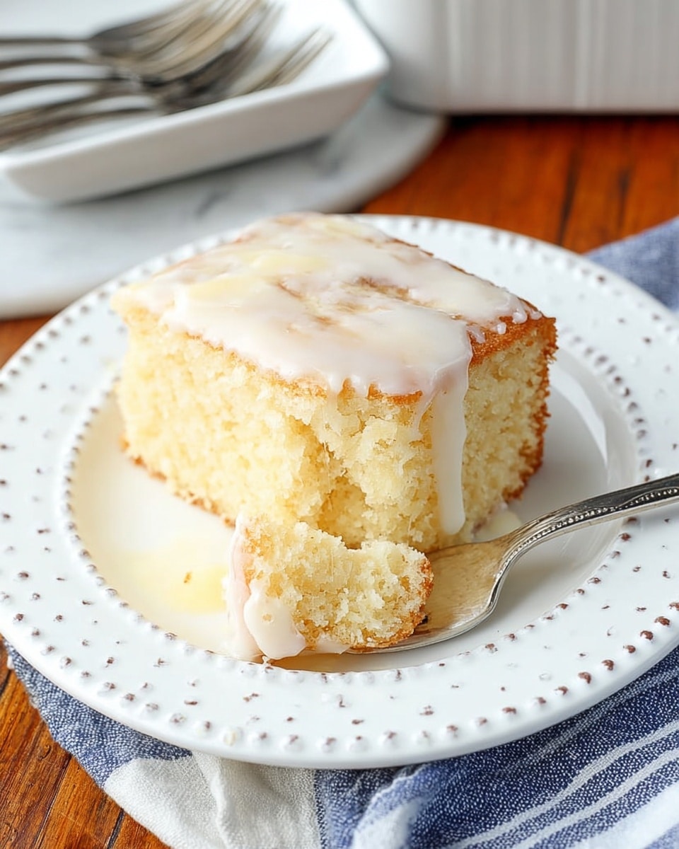 A thick square slice of soft, pale yellow cake sits on a white plate with a dotted edge, covered with a smooth white glaze that drips slightly over the sides. The cake has a moist texture with visible tiny air bubbles and some light brown swirls on top and inside. A silver fork rests on the plate, holding a small bite of the cake. The plate is placed on a wooden table with a white marbled surface visible at the edges, next to a folded blue and white striped cloth. In the top part of the image, a white rectangular dish and three silver forks are slightly out of focus. Photo taken with an iphone --ar 4:5 --v 7