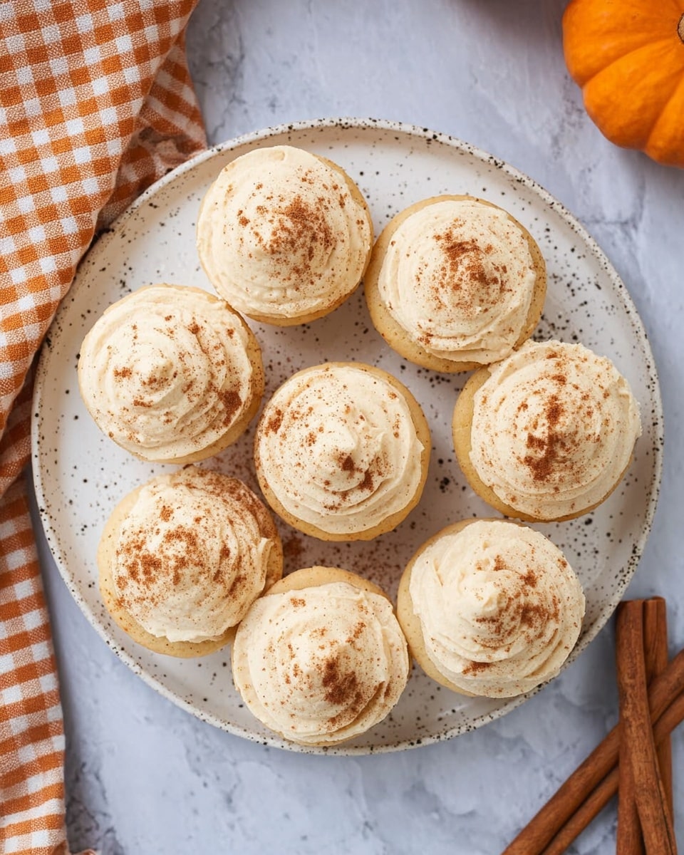 Seven light beige frosted cupcakes are arranged on a round white speckled plate with a few darker brown specks. Each cupcake has a single thick layer of smooth, swirled frosting with a slightly rough texture, topped with a light dusting of brown cinnamon powder. The plate rests on a white marbled surface with a soft mix of gray and white, next to two cinnamon sticks and an orange pumpkin partially visible in the top right corner, along with part of a checkered orange and white cloth on the top left side. photo taken with an iphone --ar 4:5 --v 7