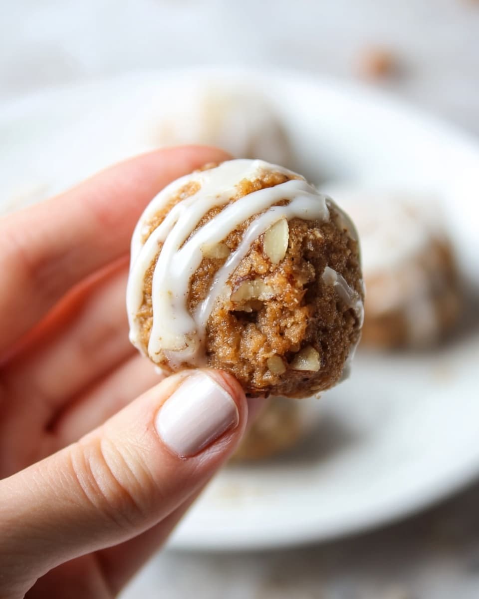 Three round cookie balls with a light brown, crumbly texture sit on a white plate. Each ball is drizzled with thin white icing lines crossing over the top. One cookie ball is partially eaten, showing a soft, crumbly inside with small pieces of nuts. The plate rests on a white marbled surface. photo taken with an iphone --ar 4:5 --v 7