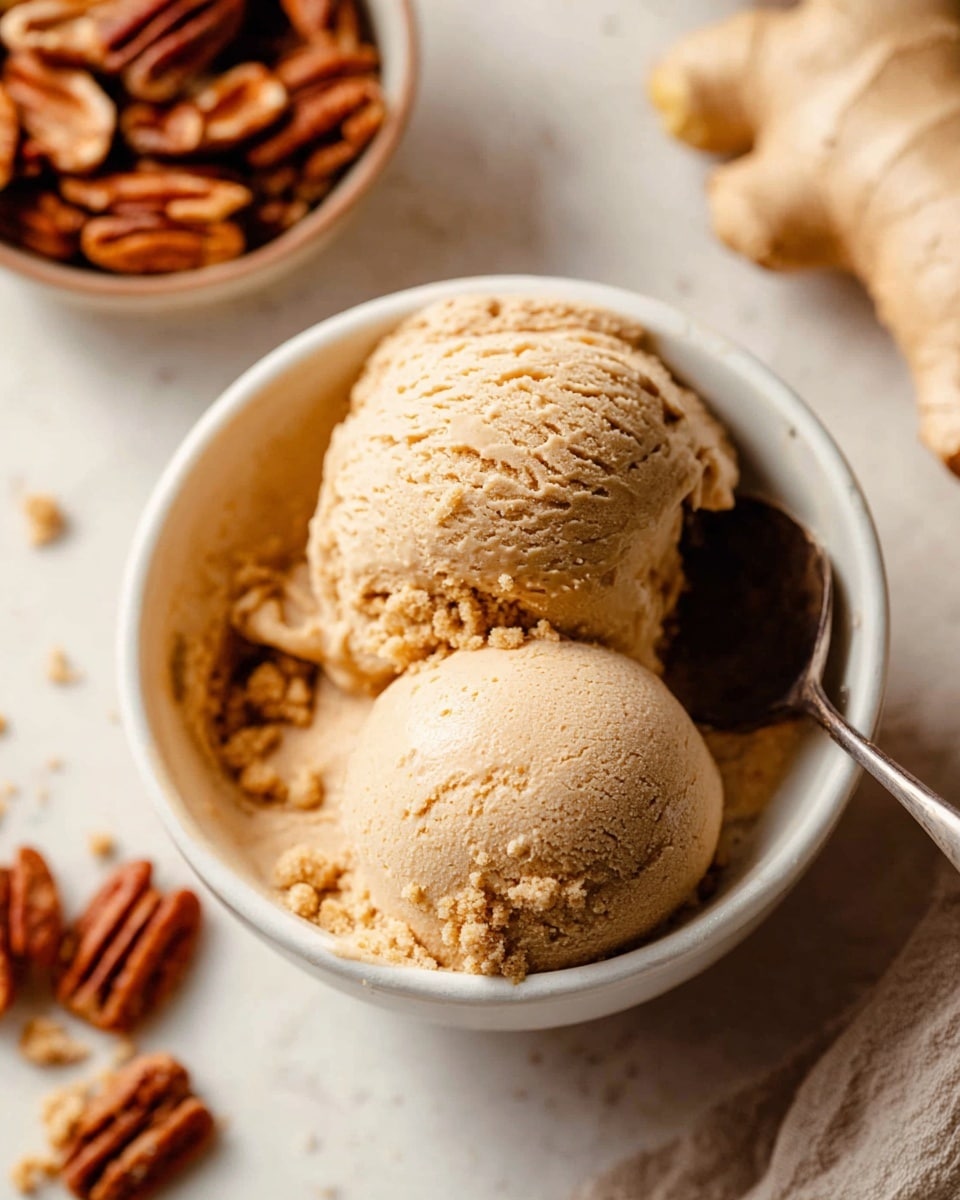 A white bowl filled with two scoops of light brown ice cream, textured with small crumbly bits on the surface, placed on a spoon inside the bowl. Around the bowl, there are some pecans in a container, and a piece of fresh ginger and a pecan are visible on the white marbled texture background. The scene has a warm and cozy feel with soft light. photo taken with an iphone --ar 4:5 --v 7