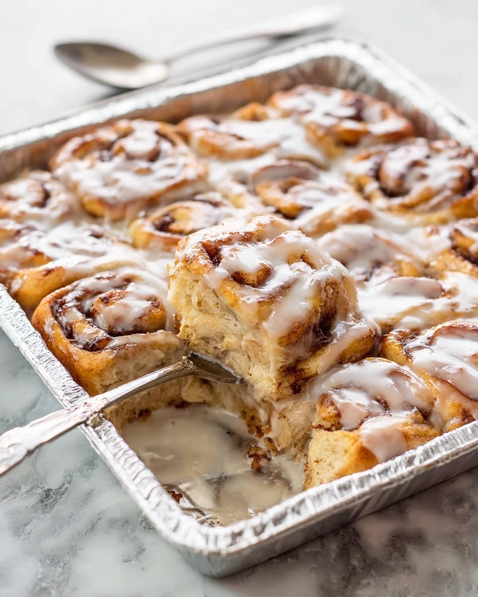A square metal baking pan lined with foil holds a cinnamon roll casserole covered in thick white glaze. The casserole is made of bite-sized pieces of golden brown cinnamon roll dough, with the cinnamon swirls visible on each piece, arranged closely together in a single thick layer. One piece is lifted on a silver fork resting inside the pan, showing the soft, doughy texture inside with hints of melted cinnamon sugar. The pan sits on a white marbled textured surface, with a silver spoon placed above it in the background. photo taken with an iphone --ar 4:5 --v 7