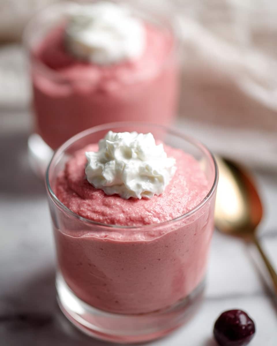 Two clear glass cups are filled with a thick, creamy pink mousse with a smooth but slightly textured surface. Each cup is topped with a small dollop of white whipped cream centered on the mousse. The cups are placed on a white marbled surface, and a blurred, shiny gold spoon lies nearby along with a small dark berry. The focus is sharp on the front cup, showing details of the mousse texture and whipped cream. photo taken with an iphone --ar 4:5 --v 7