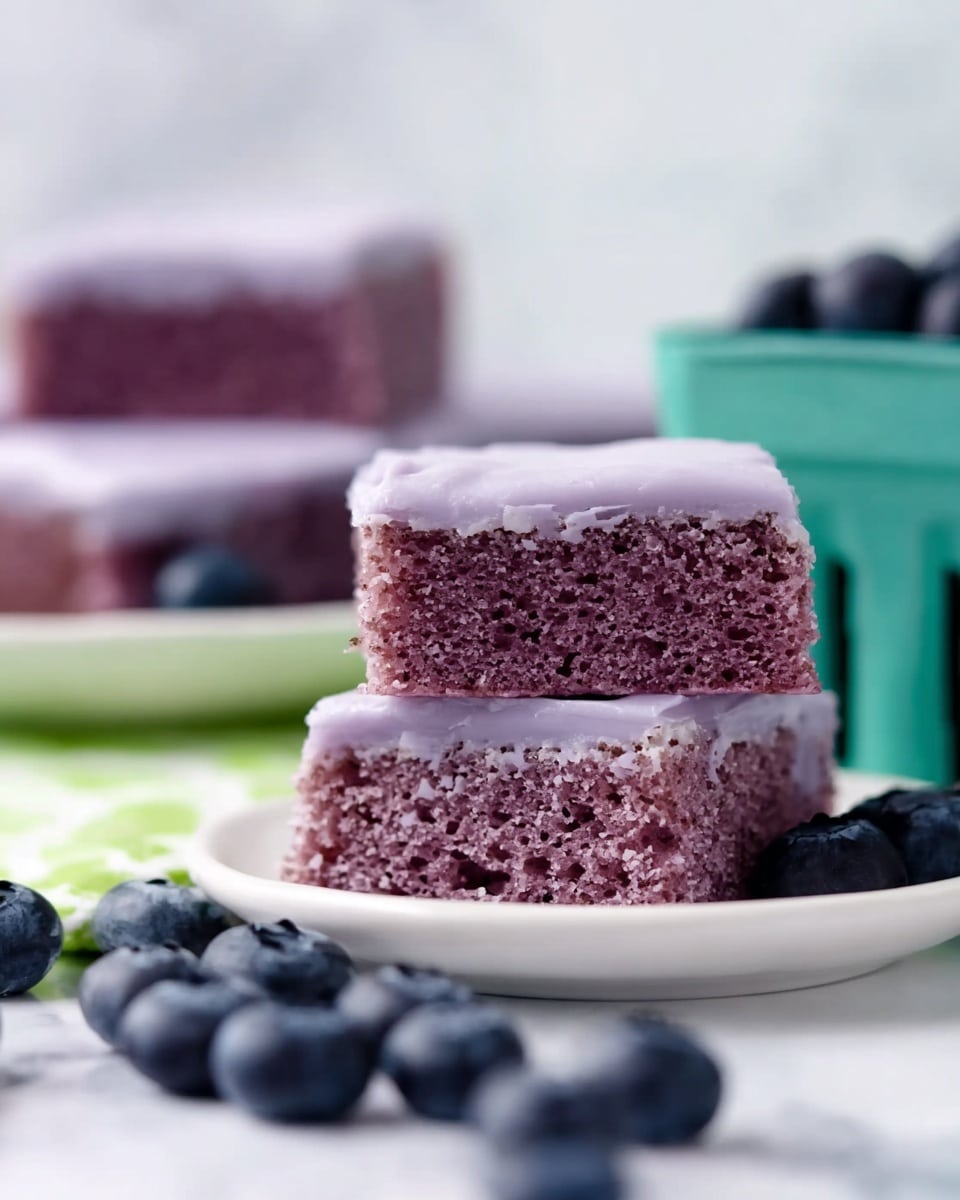 Two square purple cake pieces stacked on top of each other, each with a smooth, pale purple icing layer on top. The cake texture looks moist and dense with tiny air holes. In the background, more cake pieces are on a white plate, with scattered fresh blueberries and a green container holding more blueberries nearby. The scene rests on a white marbled texture surface. photo taken with an iphone --ar 4:5 --v 7