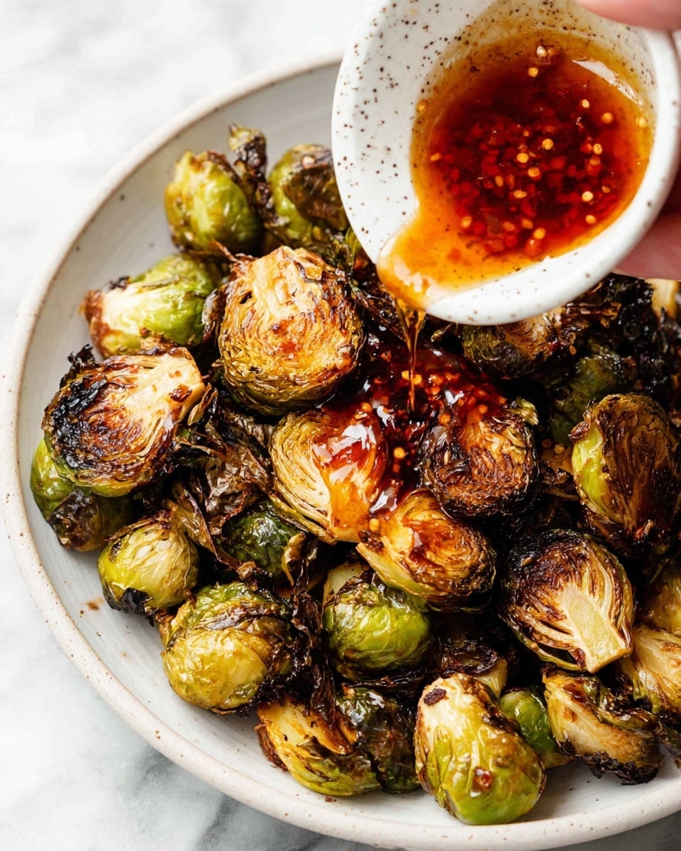 A close-up of a white bowl filled with roasted Brussels sprouts that are golden brown with some blackened crispy edges. A speckled white sauce dish is held above the bowl by a woman's hand, and a deep red chili oil sauce with chili flakes is being poured over the Brussels sprouts, glistening as it coats them. The white marbled surface is underneath the bowl. photo taken with an iphone --ar 4:5 --v 7