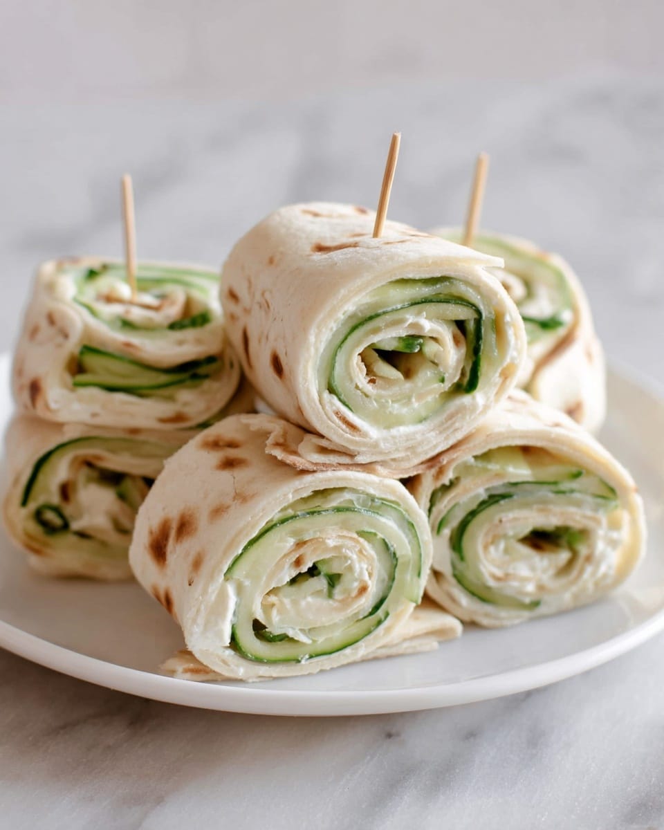 The image shows a stack of five pinwheel sandwiches on a white plate placed on a white marbled surface. Each sandwich is made of a thin flatbread rolled tightly in three visible layers; the outer layer is a light beige, soft flatbread with slight brown spots, the middle layer is a creamy white spread, and the inner layer consists of thin green cucumber slices that add a fresh texture. The sandwiches are secured with small wooden toothpicks and stacked with three on the bottom and two on top, showing a spiral pattern. photo taken with an iphone --ar 4:5 --v 7