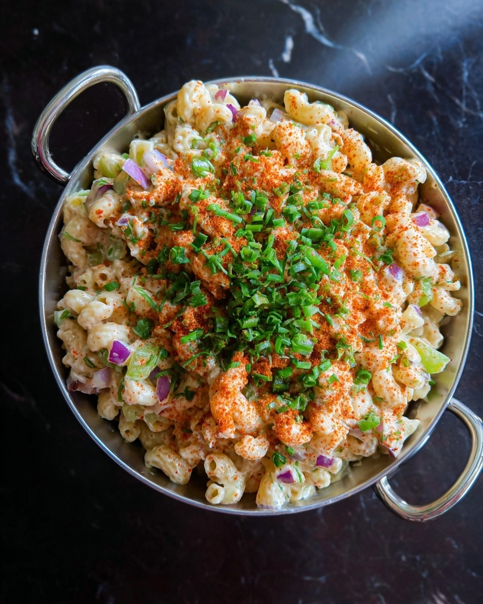 A close-up view of a large, round metal bowl filled with a mixture of cooked elbow macaroni pasta coated in a creamy, pale yellow dressing. Scattered evenly throughout are small pieces of chopped red onion with vibrant purple hues and bits of green celery, adding color contrast and texture. The pasta appears soft and slightly chunky, with some small bits of white egg visible, creating a layered, mixed look. The bowl is placed on a white marbled surface, and the handles of the bowl are visible on either side. photo taken with an iphone --ar 4:5 --v 7
