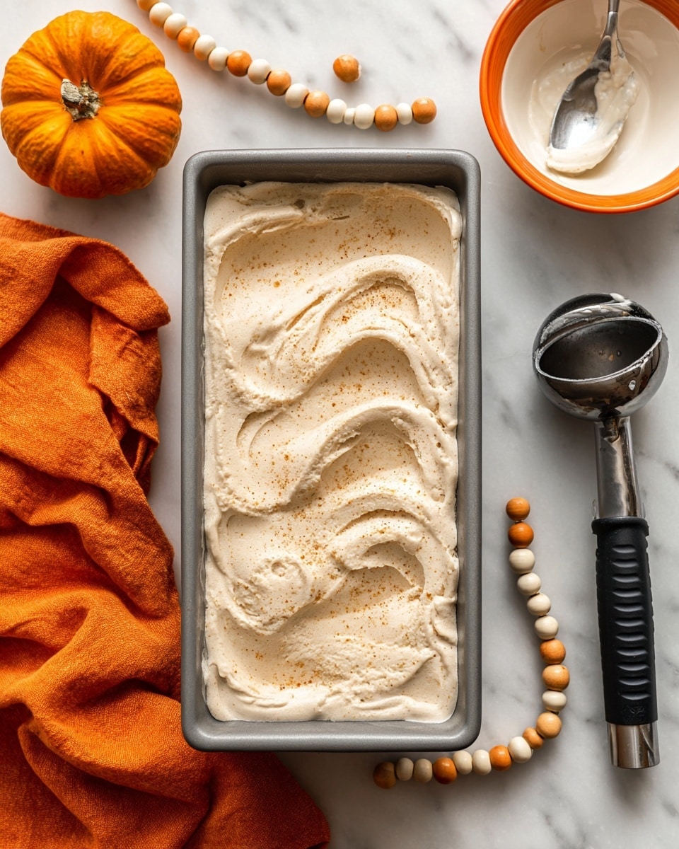 A rectangular gray loaf pan filled with a thick, creamy, light beige ice cream that has a swirled and slightly textured surface sprinkled with fine specks of spice, positioned on a white marbled countertop. To the left of the pan is a small orange pumpkin resting on a crumpled orange cloth. To the right of the pan, there is a white bowl with an orange rim and a silver spoon inside, next to a black ice cream scoop with a shiny metal head lying flat, and below them is a string of round beads in white, orange, and natural wood colors. photo taken with an iphone --ar 4:5 --v 7