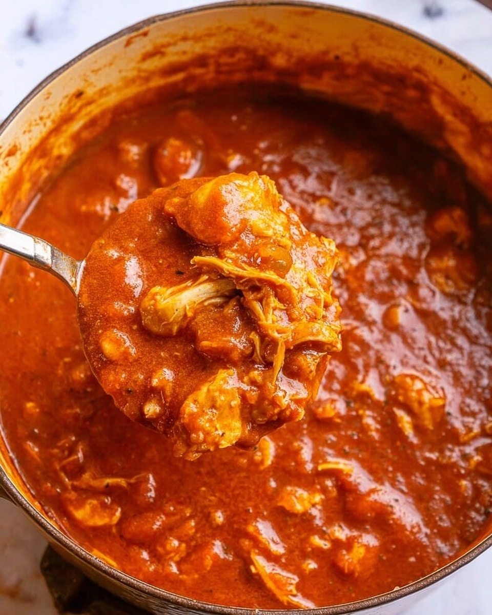 A close-up image of a pot filled with thick, rich tomato-based stew with chunks of chicken. The stew has a deep red-orange color with visible pieces of tender, shredded chicken and small bits of vegetables or seasoning throughout. A spoon is lifting a portion of the stew, showing the texture and layers of the sauce clinging to the juicy chicken. The pot's inside rim is visible, and the inside is slightly stained from the sauce. The background is a white marbled texture. photo taken with an iphone --ar 4:5 --v 7