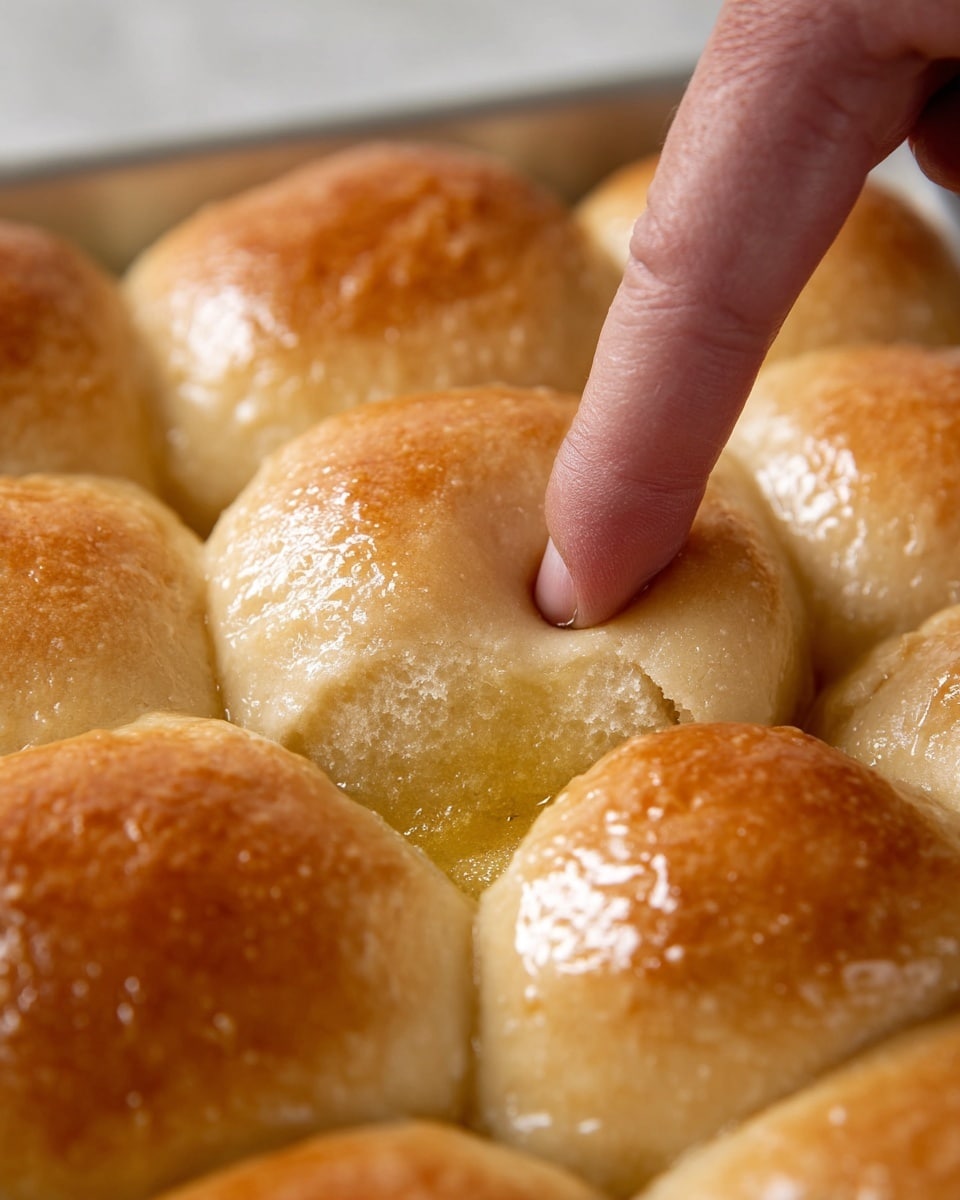 A close-up view shows a tray of soft, golden-brown dough buns with a smooth, slightly shiny surface from a butter glaze. One bun is gently pressed down in the center by a finger, creating a small dent and showing the buns' fluffy texture. The buns are placed close together, forming a neat pattern of rounded shapes, all sitting on a white marbled texture background. Photo taken with an iphone --ar 4:5 --v 7