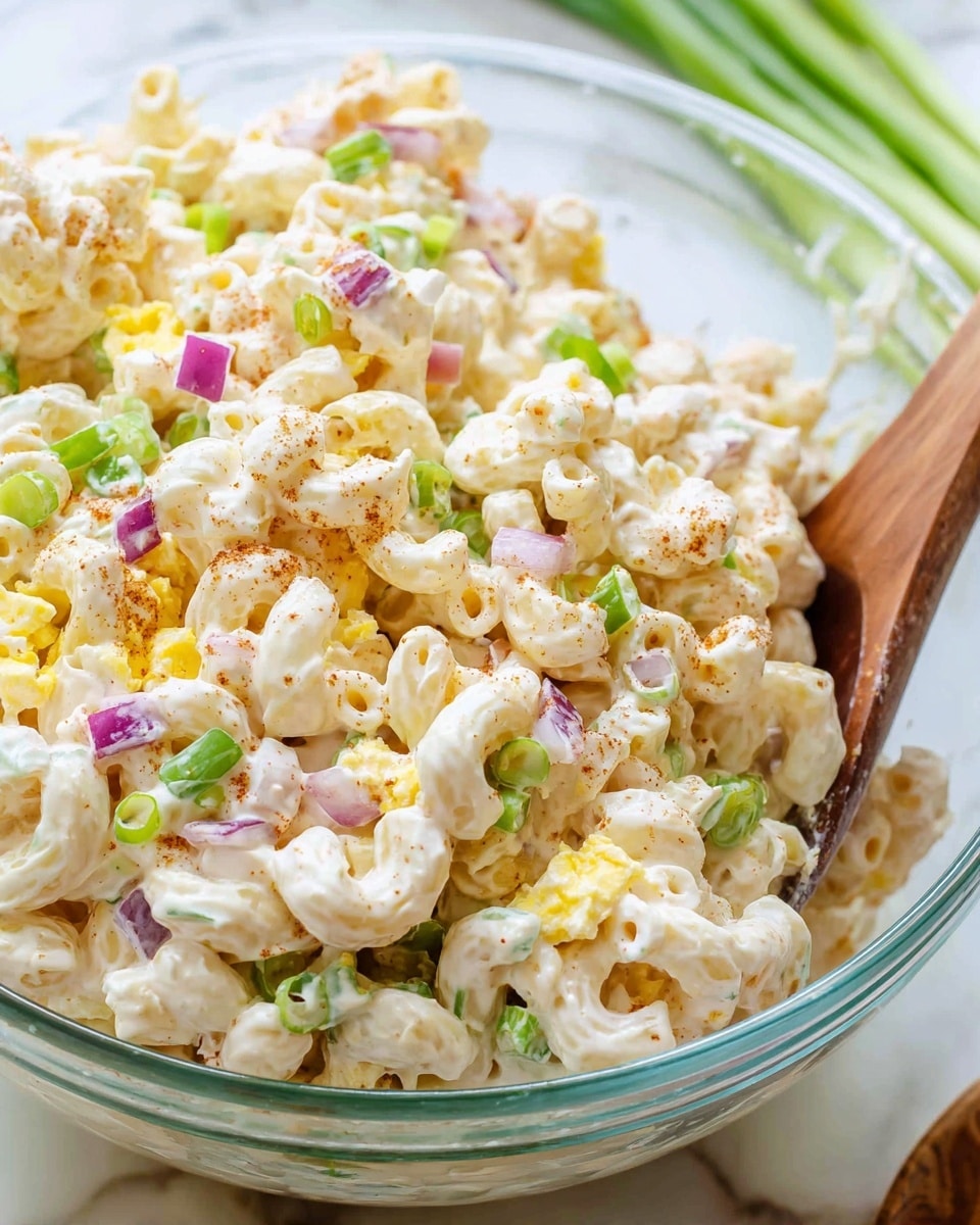 The image shows a close-up of a creamy macaroni salad in a clear glass bowl. The salad has three main layers: small, curved macaroni pasta coated in a white, thick creamy dressing forms the base, speckled with light brown spices. Scattered evenly throughout are small pieces of chopped green onions, purple-red onion bits, and yellow egg chunks, adding a mix of green, purple, and yellow colors to the mix. A wooden spoon is partially visible on the right side, mixing the ingredients. The bowl is placed on a white marbled surface with some green onion pieces blurred in the background. Photo taken with an iphone --ar 4:5 --v 7