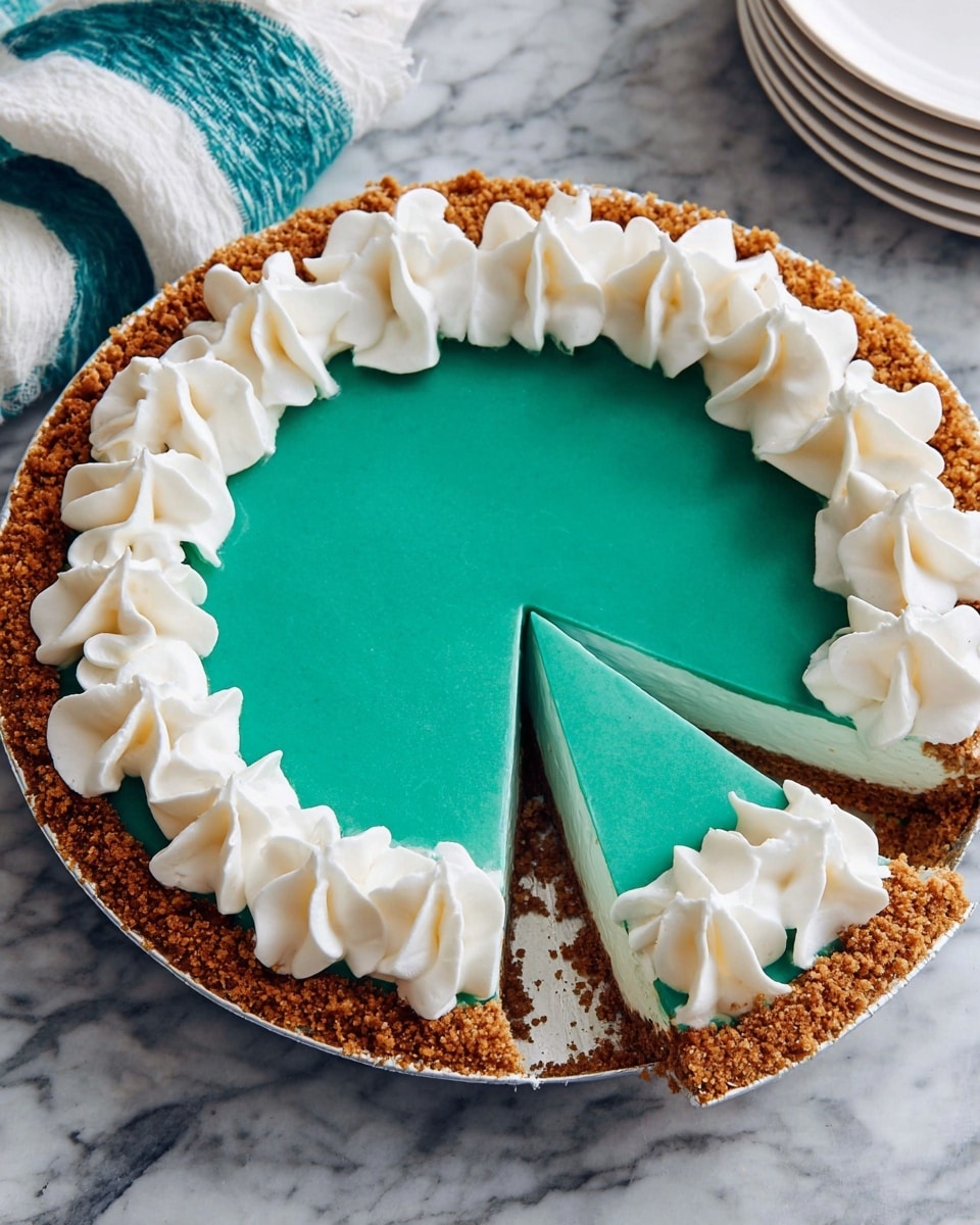 A round pie with a thick light brown crumb crust lines the outer edge evenly; inside is a smooth, shiny bright teal filling that forms the main layer. The top edge of the pie is decorated with large dollops of white whipped cream forming a ring around the entire circle. A single slice is cut out from the bottom, showing the clear separation of the creamy teal filling from the crust. The pie is on a metal pie pan sitting on a white marbled textured surface, with a folded white and teal cloth in the top left corner and a stack of white plates in the top right corner. photo taken with an iphone --ar 4:5 --v 7