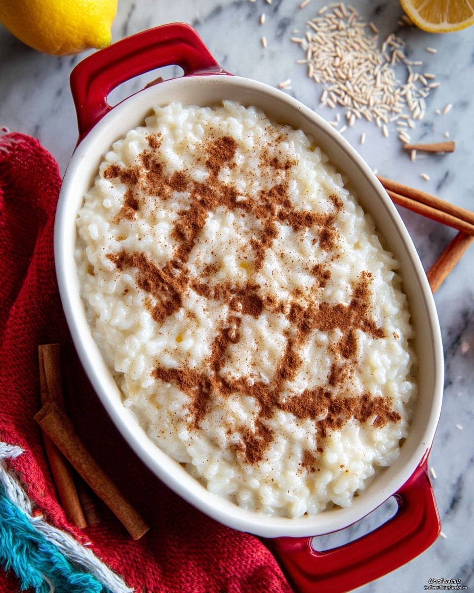An oval white dish with red handles filled with creamy white rice pudding, topped with cinnamon powder in a crisscross pattern creating diamond shapes. The rice pudding has a smooth texture with visible soft rice grains throughout. The dish is placed on a white marbled surface with a red and white cloth below, a cinnamon stick and some loose rice grains scattered nearby, and a yellow lemon in the background. photo taken with an iphone --ar 4:5 --v 7