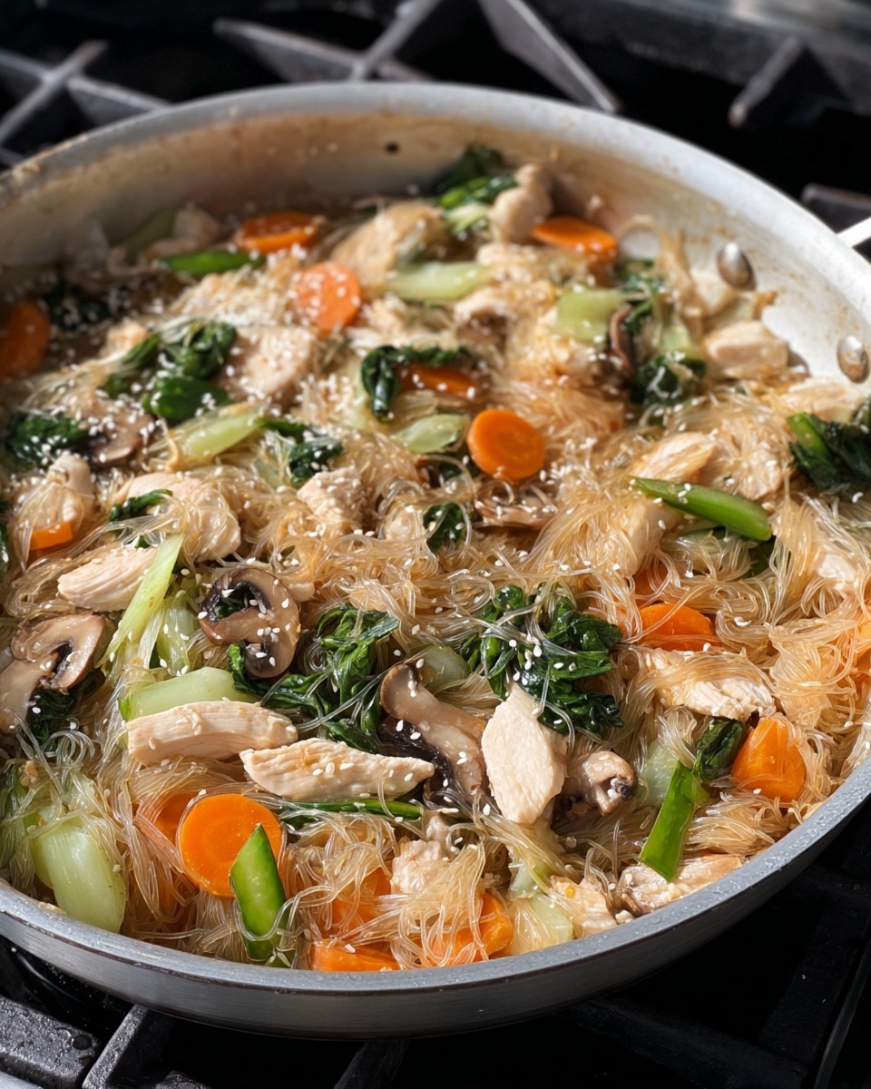 A close-up view of a light gray pan filled with a stir-fry dish showing three main layers: translucent thin noodles forming the base with a glossy texture, tender white chicken slices scattered over the noodles, and mixed vegetables including bright orange carrot slices, light green bok choy pieces, dark green leafy greens, and small brown mushroom slices spread evenly on top. White sesame seeds are sprinkled on the dish, adding small, light-colored specks throughout. The pan is on a stove with black grates visible in the background. photo taken with an iphone --ar 4:5 --v 7