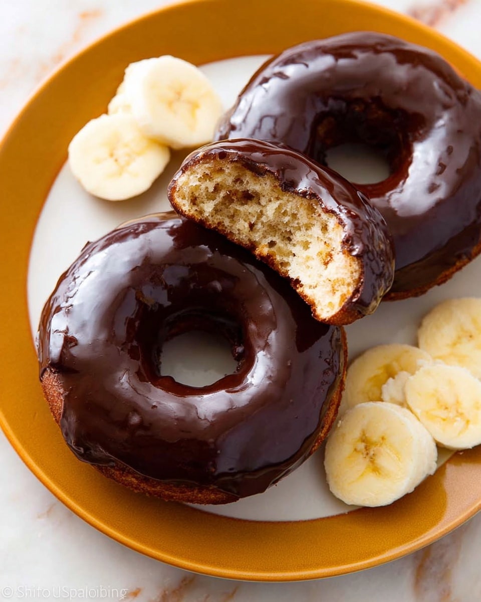 Two chocolate-glazed donuts are placed on a white plate with an orange rim set on a white marbled surface. One donut is whole with a shiny, smooth dark chocolate glaze on top. The other donut is broken into two pieces showing a light brown, soft, and fluffy cake inside beneath the glossy chocolate glaze. Next to the donuts, there are a few slices of peeled banana with a creamy, pale yellow color and soft texture. Photo taken with an iphone --ar 4:5 --v 7
