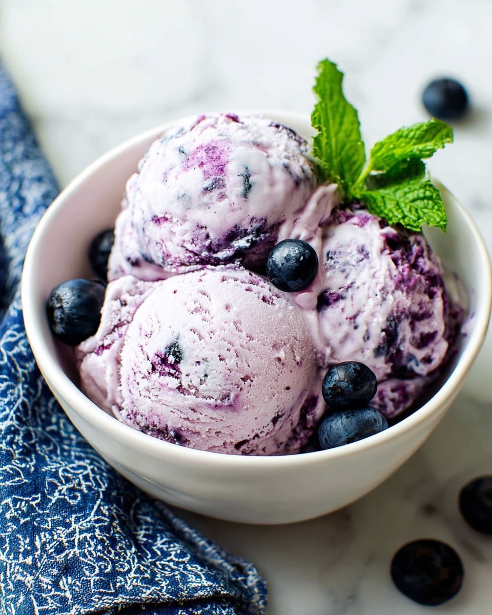 A white bowl filled with three scoops of light purple ice cream mixed with darker purple berry swirls, giving it a creamy and textured look. Around the scoops are several dark blue blueberries, and a fresh green mint leaf sits on top for contrast. The bowl is placed on a white marbled surface, with a blue patterned cloth nearby, creating a fresh and inviting scene. Photo taken with an iphone --ar 4:5 --v 7