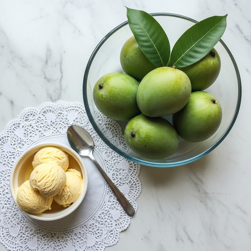 The image shows a round clear glass bowl filled with six light green mangoes and three large green leaves on top, placed on a white marbled surface. To the left of the bowl, there is a small white bowl containing three scoops of pale yellow ice cream, placed on a white lace doily with intricate patterns. A silver spoon rests near the ice cream bowl, partly on the doily. The overall setting contrasts the smooth textures of the glass, fruit, and ice cream with the delicate lace and the white marbled background. photo taken with an iphone --ar 4:5 --v 7