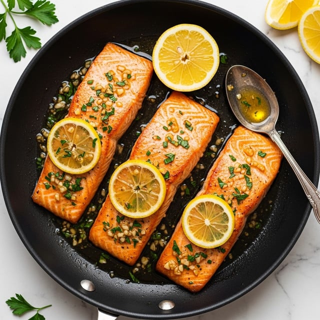 Three cooked salmon fillets with a golden brown surface sit side by side in a black pan. Each fillet has a shiny glaze of oil with small bits of minced garlic and sprinkled green herbs on top. Thin yellow lemon slices rest on two of the fillets, while one lemon wedge lies beside the salmon in the pan. A silver spoon with some glaze on the bowl is placed near the right edge inside the pan. The pan is set on a white marbled surface, with some fresh parsley and extra lemon slices nearby. Photo taken with an iphone --ar 4:5 --v 7