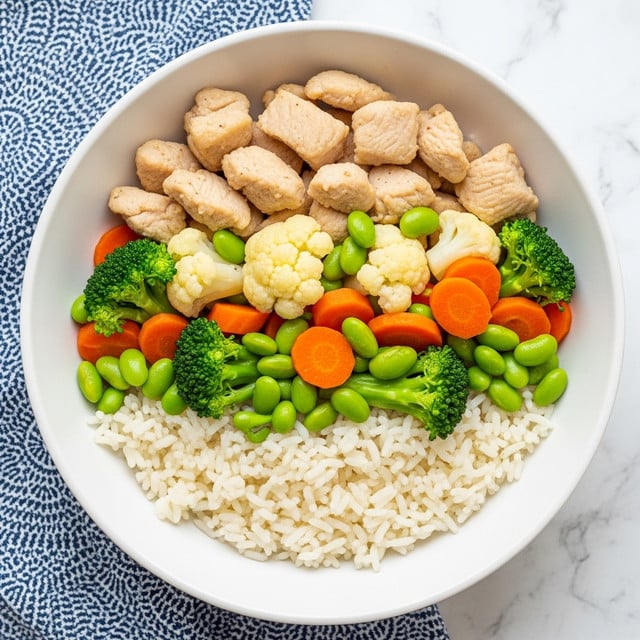 A white rectangular plate filled with a colorful stir-fry dish layers tender light brown chicken pieces mixed with bright orange carrot slices, light green broccoli florets, pale cauliflower chunks, and vibrant green edamame beans. Thin almond slices are scattered on top, adding texture with their pale tan color. The food looks glossy and well-cooked. Behind the plate, there is a white bowl filled with fluffy white rice, sitting on a bright green cloth. A metal spoon lies to the side on a white marbled surface that replaces the wooden table. Photo taken with an iphone --ar 4:5 --v 7